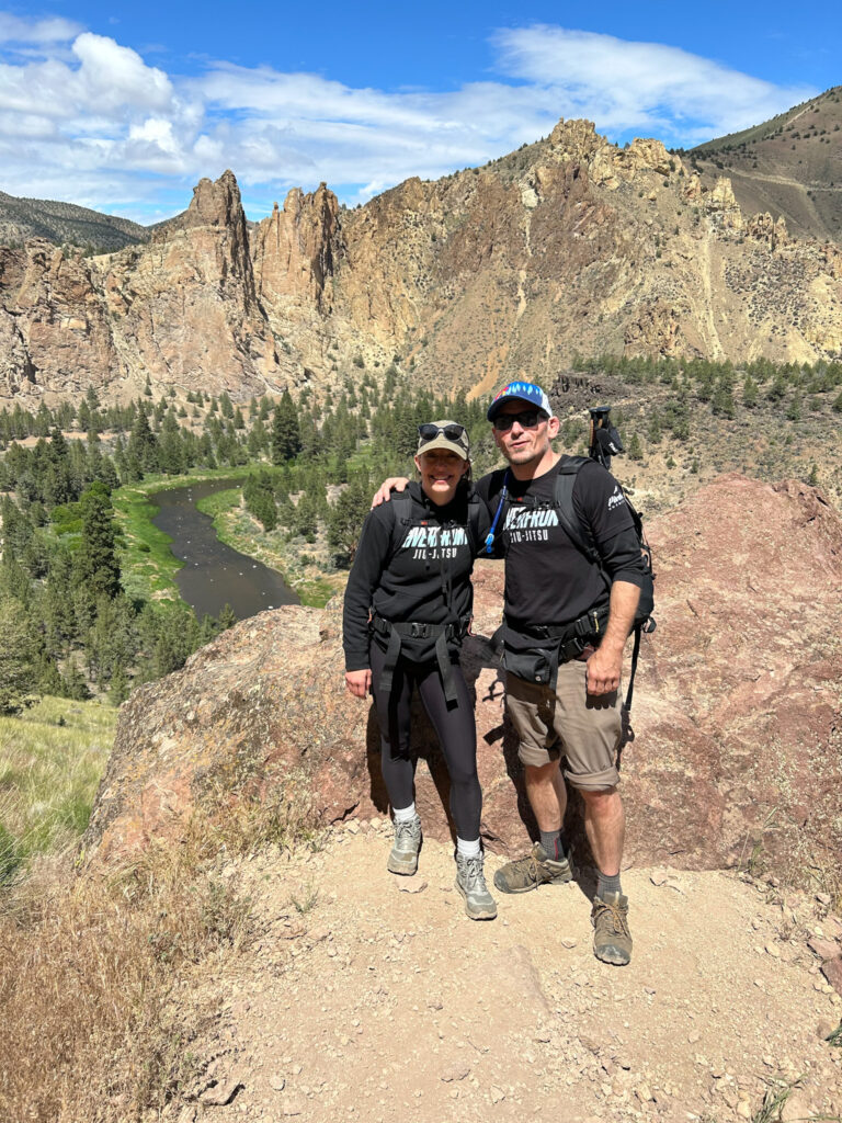 A man and woman standing in front of scenic mountains at the top of a hike. 