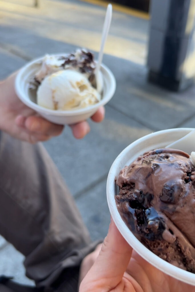 Two cups of ice cream held by two peoples hands with two scoops and a spoon sticking out of it outside of the ice cream shop.